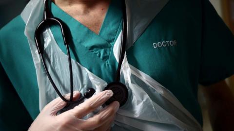 A doctor wearing green scrubs, with a plastic apron and latex gloves who is holding a black stethoscope against his chest.