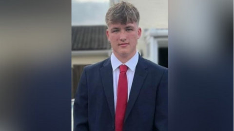Joshua Blake wearing a suit and a red tie. He is standing in front of a building and looking at the camera.