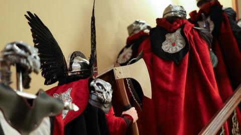 Backs of men dressed in Viking helmets and red cloaks going up stairs. One is holding an axe.