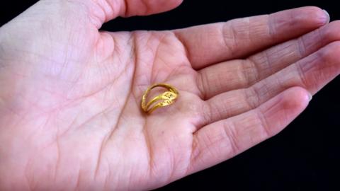 A pair of conjoined Roman gold rings, sitting in the palm of a hand. One ring is decorated with 25 tiny gold pellets arranged in a diamond shaped pattern. The other has a flat, elongated bezel decorated with fine strands of gold wire forming a central circle with drop-shaped motifs on either side, possibly representing a flower. Together, they are delicate, skilfully made objects, dating to between the 1st and 3rd centuries AD.