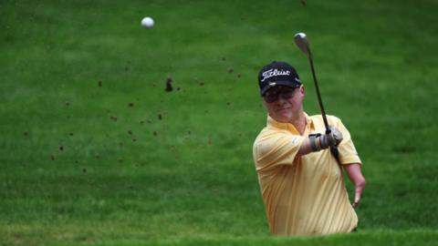 A golfer with a yellow tshirt and black cap striking the ball in the air.
