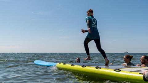 A boy jumping off a mini-jetty into the sea with volunteers around and supporting him 