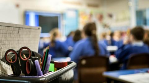 A picture of a classroom, with a box of pens, rulers and othe school equipment in focus and out of focus school pupils in the background, wearing blue uniforms