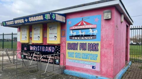 A pink ticket office, which is now closed, where people would have been able to get tickets, wristbands and change for the amusements. There is a blue sign with a carousel which reads "LEISURE ISLAND FAMILY FUN PARK, BOOK YOUR PARTY HERE".