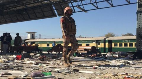 a soldier walks among debris from the bomb on a station platform with a green and yellow train in the background