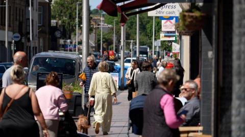A busy shopping street with numerous people sitting at pavement cafes