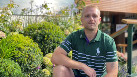 Will crouches down next to a green flowerbed. He is wearing a dark green Wimbledon polo top with white stripes across it and is looking into the camera. He has blonde hair cut short. 
