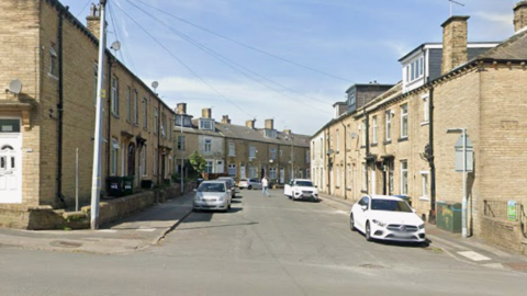 Google Street View image of a residential street. The houses are made from light brick and are all terraced. There are some cars parked either side of the road.