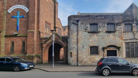 A church building with two cars parked in front of it. Part of the building is a red brick style, with a cross and The Mackintosh Halls on the wall. 