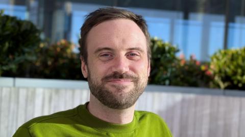 Reporter Matthew Bassett smiles into the camera. He has short, cropped dark hair and a beard and is wearing lime green jumper.