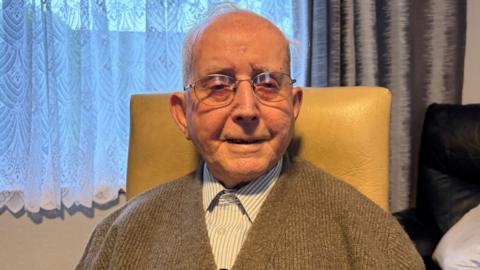 Charlie O'Leary, who is 102, sits in his living room in a tan-coloured chair. He is wearing a blue and white striped shirt buttoned up to the collar with a light brown cardigan over the top. He has round glasses on. Over his left shoulder a black leather sofa can be seen with silver pillows on it. Behind him is a window with a net curtain covering it.