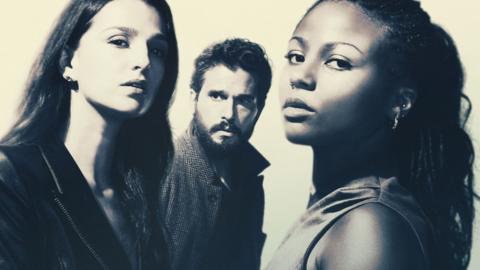 Two women stand opposite each other and look towards the camera. Behind them stands a bearded man