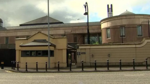 Newry Crown Court pictured which is a brown building with a cream wall outside of it, black railings on top of the wall and a black gate.