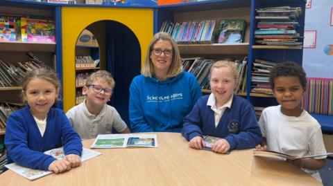 The photo shows four schoolchildren, and one woman smiling at the camera. The pupils are wearing school uniform, with a boy and girl sitting on either side of the adult. The woman in the middle is wearing a bright blue hoodie with the word 'Schoolreaders' written on it. They are all sitting at a table with books in front of them. There are bookshelves filled with books behind them. 