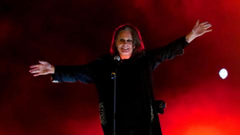 A man stands in front of a microphone against a red stage backdrop. He has chin-length dark hair and he is wearing a black long-sleeved tunic. He has black eyeliner around his eyes.