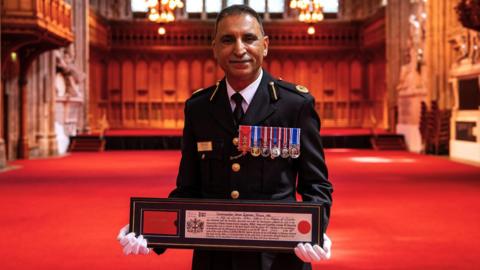 Umer Khan stands in traditional uniform with medals on display, holding up a certificate.