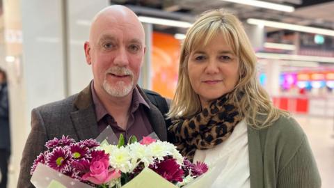 A man with a white goatee beard holding a big bunch of flowers, standing next to a blonde haired woman wearing a white top and green cardigan