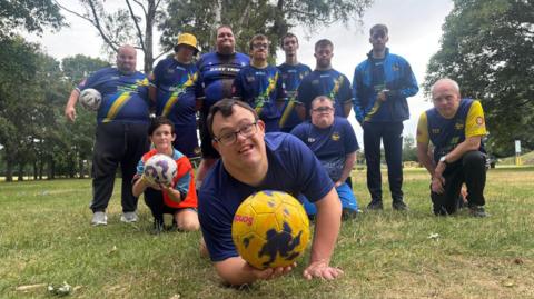 Group portrait of a community football team on a grassy park field surrounded by trees. Several participants wearing blue team kits pose together, while one participant in the foreground holds a yellow football toward the camera. Another participant kneels holding a white and purple football, with the rest of the team standing or kneeling behind.