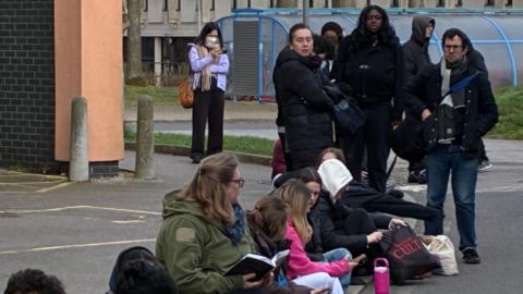 A queue of people outdoors some sat and some standing with some wearing masks