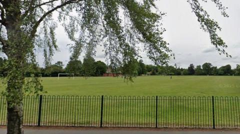 King George V playing fields in Haltemprice. The green playing fields are contained by a dark green fence. The trees are in leaf and there is a path running in front of the fence .