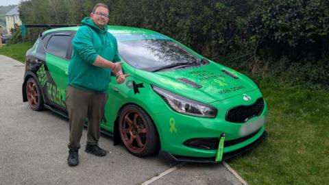 A man with brown hair and glasses and wearing a green hoodie, dark green trousers with black boots is stood in front of his bright green car. He has his thumbs up and is looking straight at the camera. 