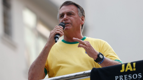 Wearing the iconic colours of the Brazilian national football team of yellow and greent, former Brazilian president Jair Bolsonaro looks towards supporters as he holds a microphone and delivers a speech.