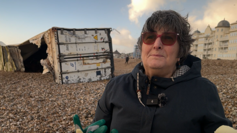 A woman with short black hair and red sunglasses stood on a pebble beach looking to the left of the camera. Behind her is a large white shipping container which is on the beach and has been badly damaged