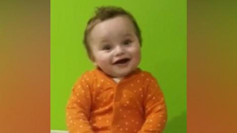 Baby in orange and white polka dot baby grow smiling while sitting on a changing table. Green wall behind him. 
