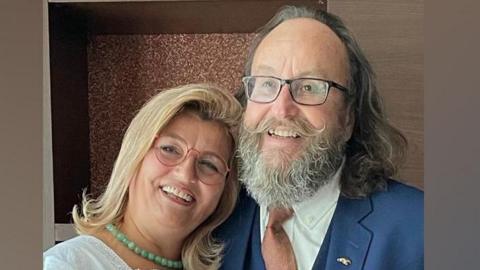 Lili and Dave Myers have their arms around each other and are smiling at the camera. Lili has mid-length blonde hair and is wearing a white top. Dave has mid-length brown hair and a grey beard. He is wearing a blue suit.