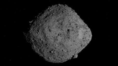 Image of a 500m-wide asteroid called Bennu that looks like a grey rock which is wider in the middle than at each end. It isn't smooth -- there are  different sized knobbly lumps sticking out from its surface. 