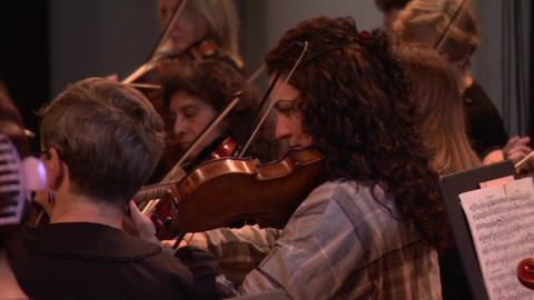 A close-up of several musicians playing violins in an orchestra, with a focus on one woman in the foreground.