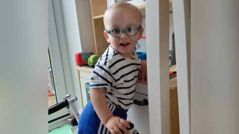 Kai, a two-year old boy, stands at a play kitchen, using a mobility aid walker. He has short blonde hair and wears blue-framed glasses, a stripy top and blue shorts.