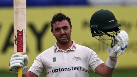 Lewis Hill celebrates reaching his century. He is raising his bat with one hand and holding his helmet aloft with the other.
