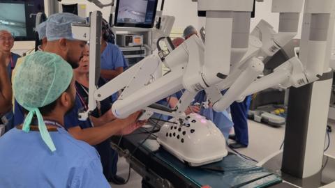 The doctors at the University Hospital of North Tees training on the Da Vinci robotic surgery machine One of the doctors, who is wearing scrubs, is operating the robotic machinery using a small control. The robotic equipment has a long surgical device inserted into a plastic shell on an operating table. A group of other doctors are watching.