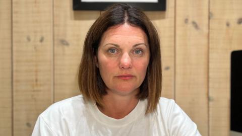 A woman with dark hair and blue eyes in a plain white T-shirt sits at a desk in a wood-panelled home office, facing the camera. A computer monitor, notebook, water bottle, phone and glasses are visible on the desk, with framed artwork hanging on the wall behind.