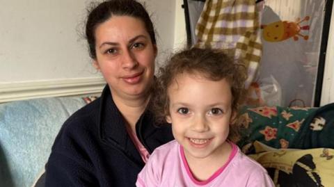 A woman with dark hair which is tied back and wearing a dark coloured fleece and a pink top has a young girl on her knee. The girl has brown curly hair which is tied back and is wearing a pink top. They are both smiling while sitting in a lounge with blankets on the sofa behind them.
