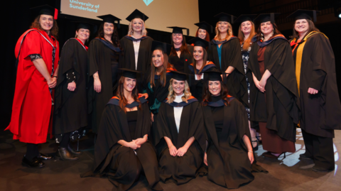 The first cohort of graduating midwives smile in their graduation black gowns and caps. They are all women of different ages. 