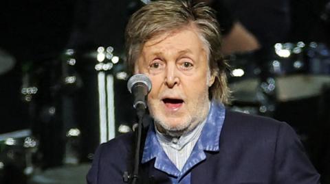 Sir Paul McCartney, with mid-length hair and wearing a blue jacket and blue shirt, sings into a microphone