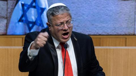 Itamar Ben-Gvir wearing a black suit, red tie and white kippah, talking on a podium in front of Israeli flags