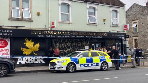 A police car parked outside an off-licence shop. There is blue and white police tape and several officers standing round outside the shop.
