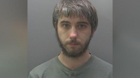 Joshua's custody image. He has black hair going grey and a black beard. He is wearing a grey t-shirt.