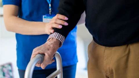 Nurs a' cumail greim air làmh seann neach a tha a' coiseachd le taic zimmer