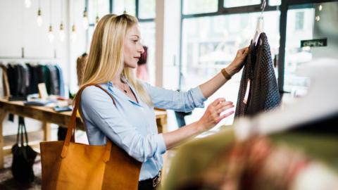 A woman shopping for clothes. She is holding up a black patterned garment on a hanger in a shop. She wears a blue blouse and has a brown bag over her shoulder as she stands in a shop in front of windows