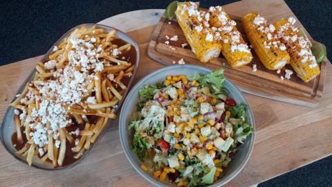 Ranch dressing in a bowl in the centre of the image, poutine on the left on a plate and corn on the cob on the right, all displayed on a wood slab. 