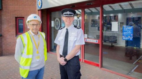 A man in a grey jumper and blue jeans is wearing a yellow lanyard, yellow hi-vis jacket and white helmet. Standing next to him is a police officer in a white shirt, black hat, with a black tie and trousers. They are standing outside a police station building with glass doors and a red entrance