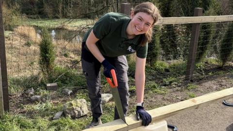 A young woman with brown hair tied back in a ponytail saws into a plank of wood. She wears a green polo shirt, black cargo trousers, working boots and gloves.