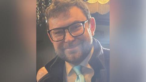 Adam Mitula with brown hair and brown beard wearing black glasses and a white shirt, pale blue tie and dark brown coat. He is smiling.
