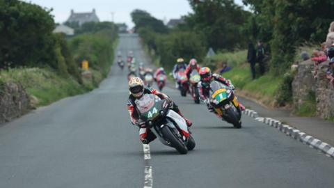 Lightweight machines on the Billown Course, which is flanked by hedgerows with some spectators seen to one side, with the leaders leaning into a right-hand bend.