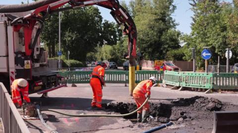 Workers in high-vis use large machinery to dig up a road while behind temporary barriers.