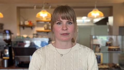A woman with blonde hair and wearing a knitted jumper. She is standing in a cafe, the counter and some lighting is visible in the background.